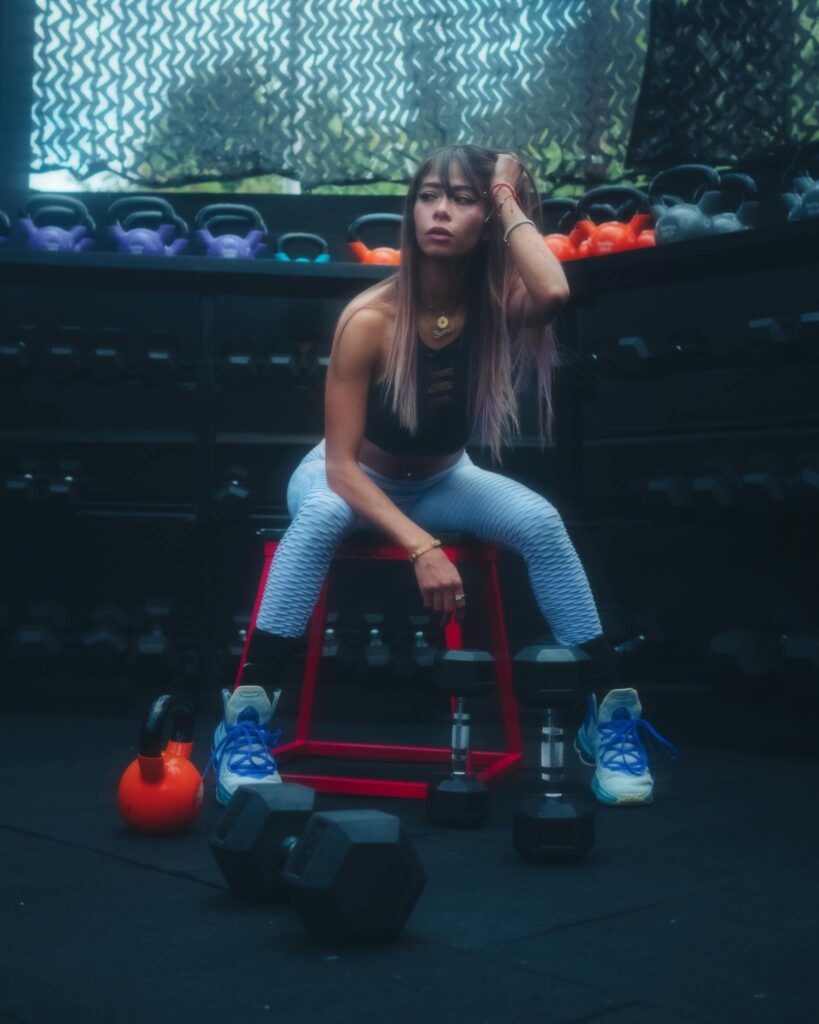 Athletic woman sitting on a stool in a gym surrounded by colorful kettlebells and weights.